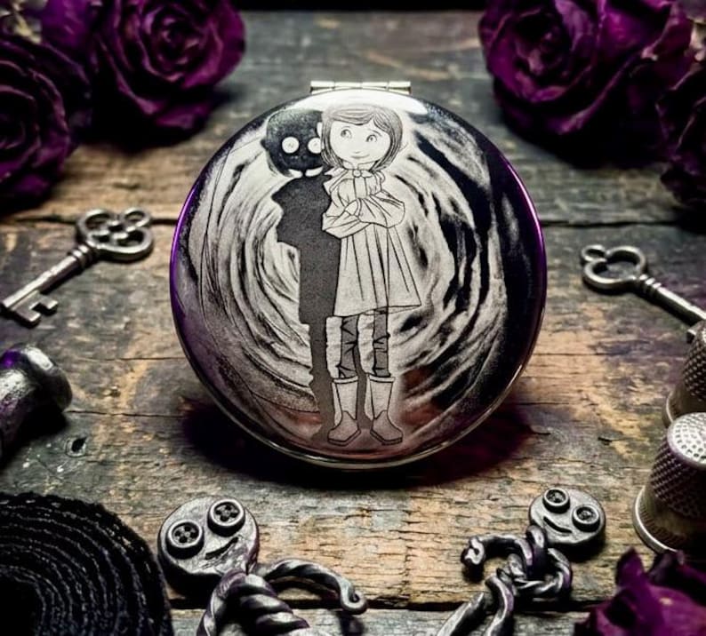 May include: A round, silver compact mirror with a detailed engraving of a girl and a shadowy figure. The design is set against a swirling background. The mirror is surrounded by keys, thimbles, and purple roses on a wooden surface.