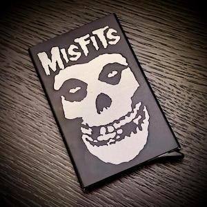 Puede incluir: Una cartera de metal negro con la palabra "Misfits" y un diseño de calavera blanca. La cartera es rectangular y está cerrada. El diseño es una imagen de alto contraste sobre un fondo oscuro.