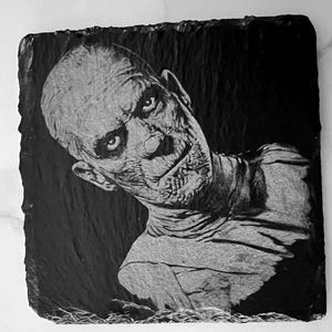 May include: Black and white image of a man's face carved into a black slate coaster. The man has a serious expression and appears to be looking to the side. The image is detailed and realistic.