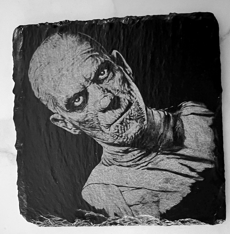 May include: Black and white image of a man's face carved into a black slate coaster. The man has a serious expression and appears to be looking to the side. The image is detailed and realistic.