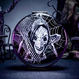May include: A round, black compact mirror with a purple rim. The mirror features a black and white illustration of a character with a sinister smile, surrounded by a spiderweb design. The image has a spooky, Halloween theme.