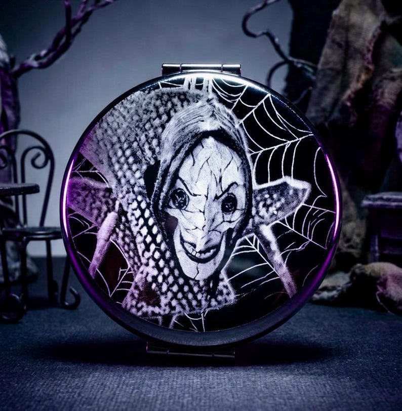 May include: A round, black compact mirror with a purple rim. The mirror features a black and white illustration of a character with a sinister smile, surrounded by a spiderweb design. The image has a spooky, Halloween theme.