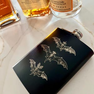 Könnte beinhalten: Schwarze Metallflasche mit silbernem Verschluss und einem gravierten Design von drei Fledermäusen. Die Flasche steht auf einer weißen Marmorfläche.