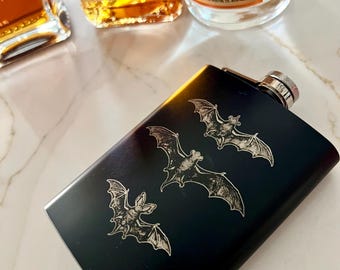 May include: Black metal flask with a silver cap and an engraved design of three bats. The flask is on a white marble surface.
