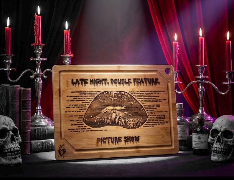 May include: A rectangular bamboo cutting board with the text "LATE NIGHT, DOUBLE FEATURE" and "PICTURE SHOW" engraved on it. The board features a detailed lip design and additional text. The scene includes candles, skulls, and bottles.