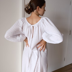 Puede incluir: Un vestido blanco de manga larga con cuello fruncido y un lazo en la espalda. El vestido está hecho de una tela ligera y texturizada.