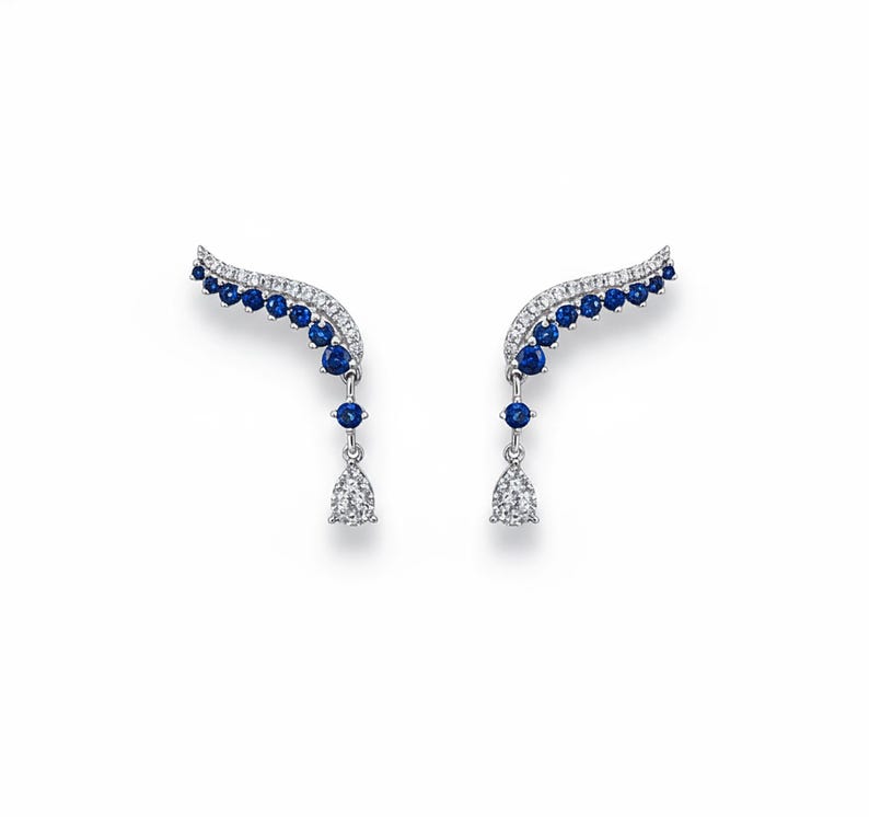 Pair of blue sapphire and diamond-style leaf ear climber earrings on a pure white background. The earrings feature a silver vine design set with round blue gemstones and a teardrop-shaped clear stone dangling at the bottom. Realistic soft drop shadows included.