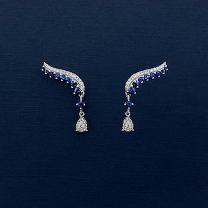 Puede incluir: Un par de pendientes trepadores plateados con un dise&ntilde;o curvo. Los pendientes presentan una fila de peque&ntilde;as piedras blancas brillantes a lo largo del borde superior, con una l&iacute;nea de piedras azules debajo. Cada pendiente tiene un colgante en forma de l&aacute;grima.