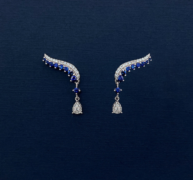 Puede incluir: Un par de pendientes trepadores plateados con un dise&ntilde;o curvo. Los pendientes presentan una fila de peque&ntilde;as piedras blancas brillantes a lo largo del borde superior, con una l&iacute;nea de piedras azules debajo. Cada pendiente tiene un colgante en forma de l&aacute;grima.