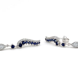 Puede incluir: Un par de pendientes elegantes con un dise&ntilde;o curvo, con una fila de peque&ntilde;as piedras preciosas de color azul oscuro y un grupo de piedras claras y brillantes. Los pendientes tienen un acento colgante en forma de l&aacute;grima.