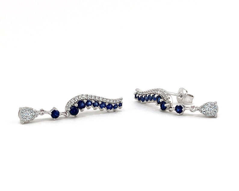 Puede incluir: Un par de pendientes elegantes con un dise&ntilde;o curvo, con una fila de peque&ntilde;as piedras preciosas de color azul oscuro y un grupo de piedras claras y brillantes. Los pendientes tienen un acento colgante en forma de l&aacute;grima.