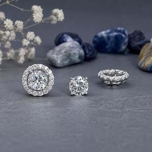 May include: Three pieces of jewelry are displayed on a gray surface. A round diamond stud earring is in the center, flanked by a halo stud earring and a diamond eternity ring. White flowers and blue stones are in the background.