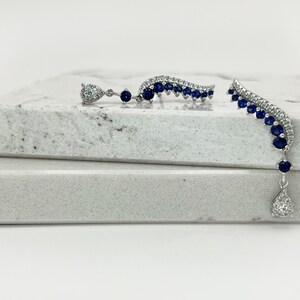 Puede incluir: Un par de pendientes plateados. Uno presenta un dise&ntilde;o curvo con piedras azules y un colgante en forma de l&aacute;grima con una gema transparente. El otro pendiente tiene un dise&ntilde;o similar. Los pendientes est&aacute;n sobre una superficie blanca.