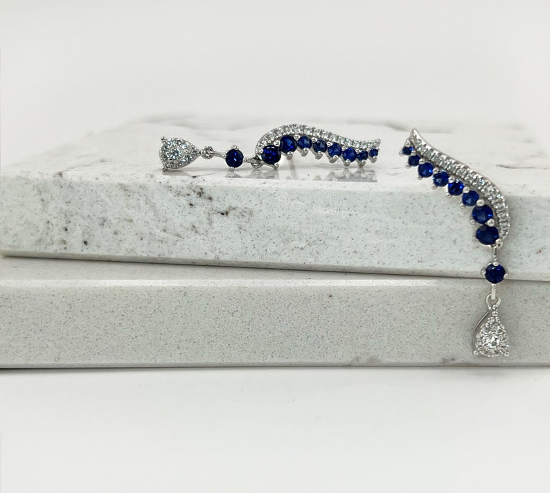 Puede incluir: Un par de pendientes plateados. Uno presenta un dise&ntilde;o curvo con piedras azules y un colgante en forma de l&aacute;grima con una gema transparente. El otro pendiente tiene un dise&ntilde;o similar. Los pendientes est&aacute;n sobre una superficie blanca.