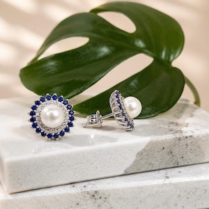Puede incluir: Pendientes de botón con una gran perla blanca central rodeada de pequeñas piedras preciosas azules, engastadas en metal plateado. Los pendientes se exhiben sobre una superficie de mármol blanco y gris, con una gran hoja verde en el fondo.