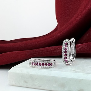 May include: A pair of silver hoop earrings with a row of small red gemstones and white gemstones around the hoop.