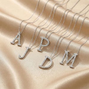 Puede incluir: Una colección de collares plateados con iniciales, cada uno con una letra diferente del alfabeto. Las letras están incrustadas con pequeñas piedras brillantes y cuelgan de delicadas cadenas. Los collares se exhiben sobre una tela beige suave.