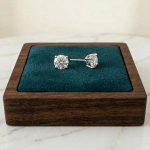 May include: A pair of diamond stud earrings displayed on a dark wooden jewelry box with a teal velvet interior. The earrings are round-cut, with a brilliant sparkle.