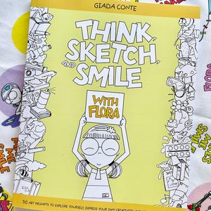 May include: A yellow book with the title "Think Sketch and Smile with Flora" and a cartoon illustration of a girl holding a sign that says "With Flora". The book has 50 art prompts to explore yourself, express your own creativity, and practice positivity.
