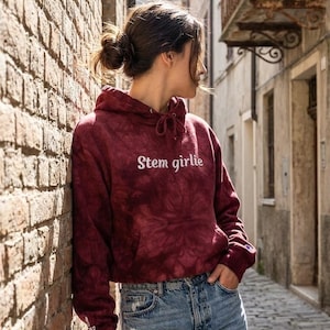May include: A maroon tie-dye hooded sweatshirt with a front kangaroo pocket and white text that reads Stem girlie. The long-sleeved pullover features a drawstring hood and a small logo patch on the left sleeve cuff, paired with light blue jeans.