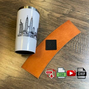 Peut inclure: Un gobelet de voyage blanc avec une illustration en noir et blanc d'un horizon urbain. Le gobelet est posé sur un dessous de verre en cuir marron avec un patch en cuir noir. Le dessous de verre est sur une surface en bois. L'image comprend également du texte pour les formats de fichiers PDF, CDR et DXF.