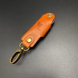 May include: A brown leather key holder with a bear silhouette and the text "HAND MADE" embossed on the front. The key holder has a brass clasp and a brass ring.