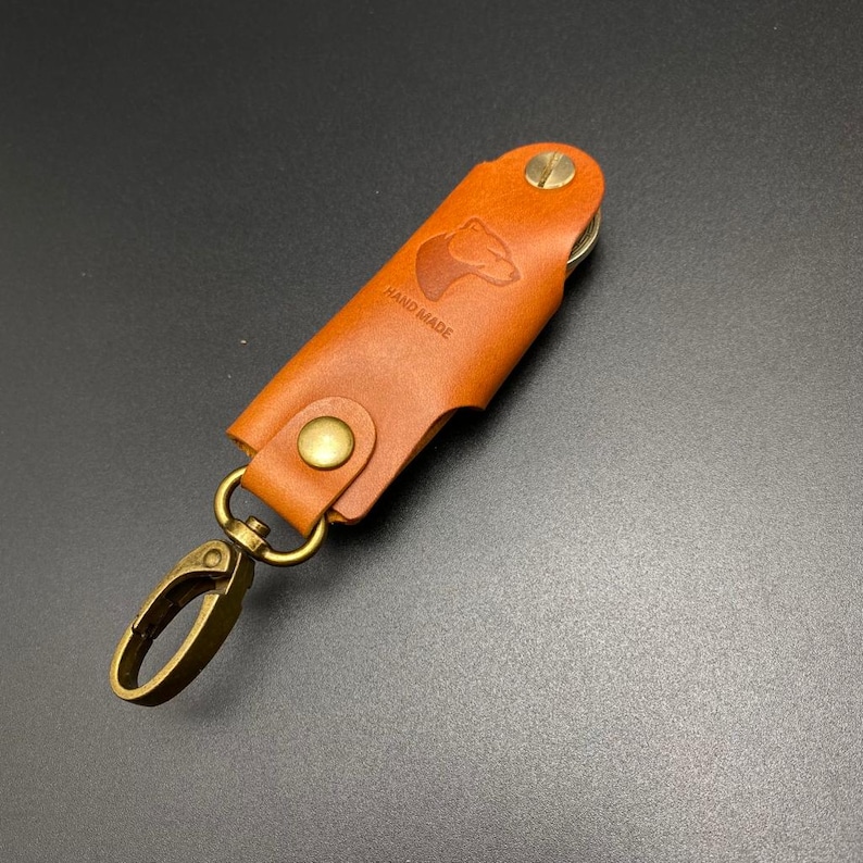 May include: A brown leather key holder with a bear silhouette and the text "HAND MADE" embossed on the front. The key holder has a brass clasp and a brass ring.