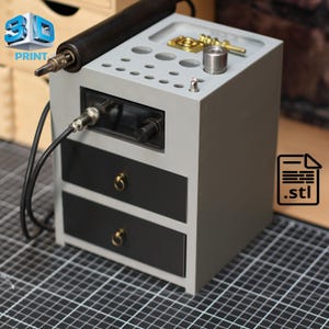 Könnte beinhalten: Eine graue 3D-Druckstation mit einem schwarzen Bedienfeld, zwei schwarzen Schubladen mit goldenen Griffen und einer Oberseite mit verschiedenen Löchern und Metallkomponenten. Ein schwarzes Kabel und ein 3D-Druckwerkzeug sind angebracht. Der Text "3D PRINT" und ein .stl-Dateisymbol sind enthalten.