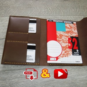 Könnte beinhalten: Eine braune Leder-Notizbuchhülle mit zwei Kartenfächern und einem Klappverschluss. Die Hülle ist geöffnet und zeigt ein Notizbuch mit einem roten und blauen Einband, auf dem "It's CREATIVE SCHOOL." steht. Das Notizbuch hat ein Rastermuster und den Text "52 kariert 5x5mm 32 Blatt".