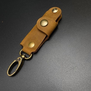 May include: A brown leather key holder with a brass key ring and brass accents. The key holder has a flap that closes with a snap.