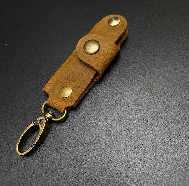 May include: A brown leather key holder with a brass key ring and brass accents. The key holder has a flap that closes with a snap.