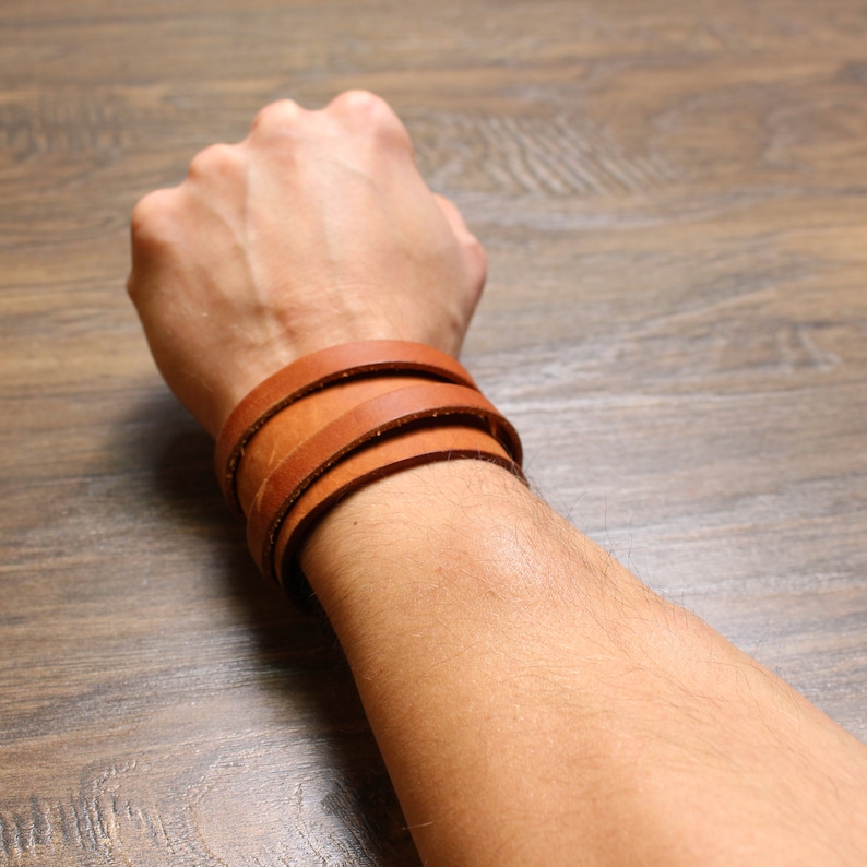 May include: A brown leather cuff bracelet with three layers of leather. The bracelet is worn on a person's wrist.
