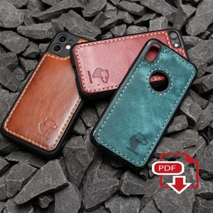 Puede incluir: Fundas de teléfono de cuero en tonos marrón, rojo y verde azulado, con costuras en contraste. Cada funda presenta un logotipo de oso en relieve y las palabras "HAND MADE". Las fundas se muestran sobre un lecho de rocas de color gris oscuro.