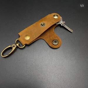 May include: A brown leather key holder with a metal clip and a key attached. The key holder has a flap that folds over the key and secures with a snap.