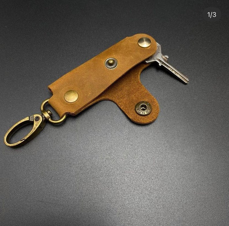 May include: A brown leather key holder with a metal clip and a key attached. The key holder has a flap that folds over the key and secures with a snap.