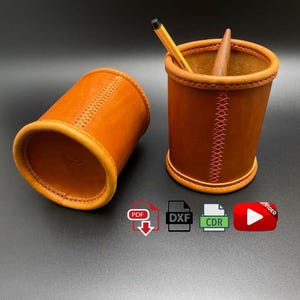 Può includere: Due portapenne in pelle marrone con cuciture rosa. Un portapenne contiene una matita e un portapenne in legno. I portapenne sono su una superficie grigia. Le icone di download PDF, DXF, CDR e video sono visibili sotto i portapenne.