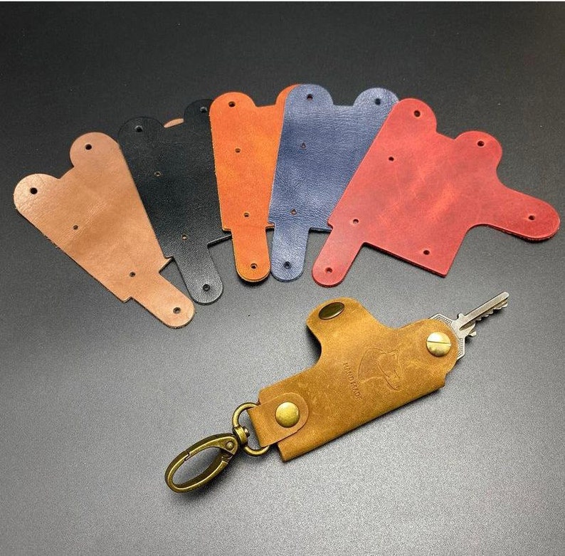 May include: Six leather keychains in different colors: brown, black, orange, blue, red, and tan. The keychains are shaped like a small, flat pouch with a hole for a key ring. A brown leather keychain with a metal key ring and a silver key is attached to the pouch.