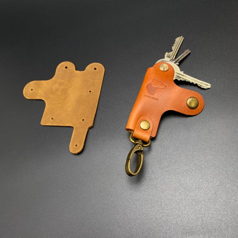 May include: A brown leather key holder with a metal ring and a small bear logo. The text "HAND MADE" is embossed on the leather. The key holder is shown next to a brown leather blank.