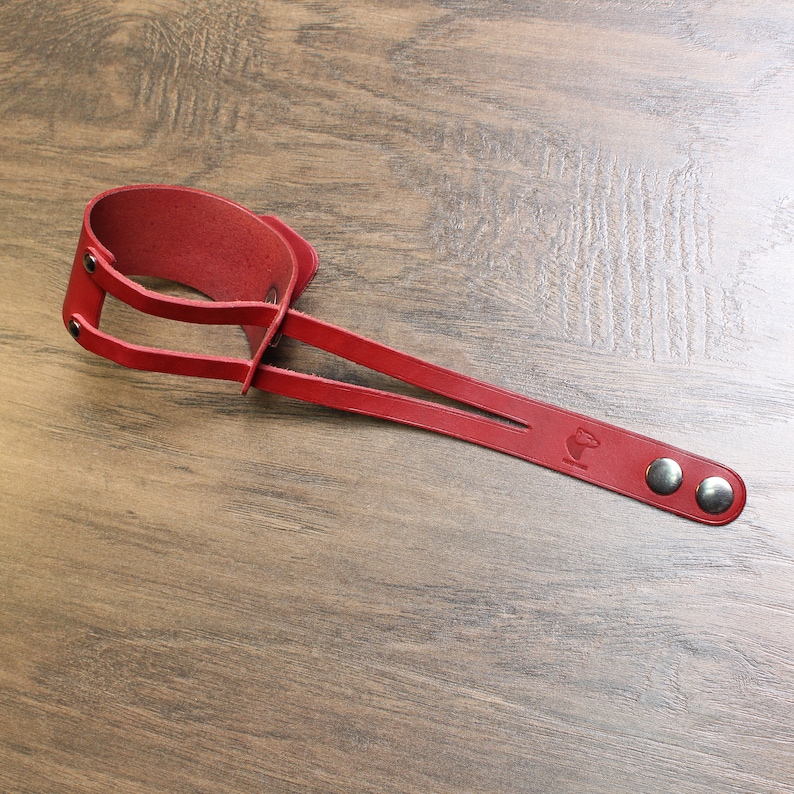 May include: A red leather wrist strap with two straps and two silver metal snaps. The strap has a small logo on it.