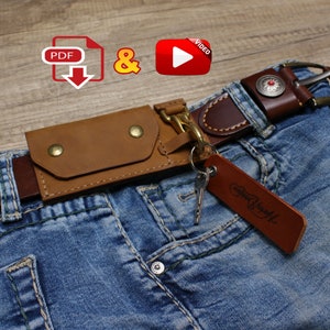 May include: A brown leather key holder with a metal ring and a key attached. The key holder is attached to a brown leather belt with a metal buckle. The belt is worn on a pair of blue jeans.