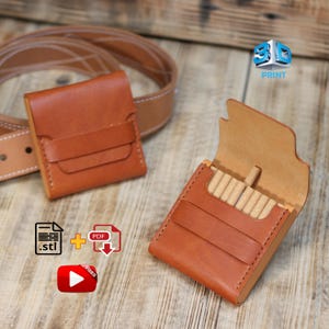 Könnte beinhalten: Zwei braune Zigarettenetuis aus Leder, eines offen und eines geschlossen, liegen auf einer Holzoberfläche. Das offene Etui zeigt Zigaretten. Ein brauner Ledergürtel ist im Hintergrund. Die Etuis sind genäht und haben eine Vordertasche.