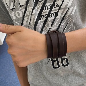 May include: A brown leather bracelet with three straps. The bracelet is worn on a person's wrist.