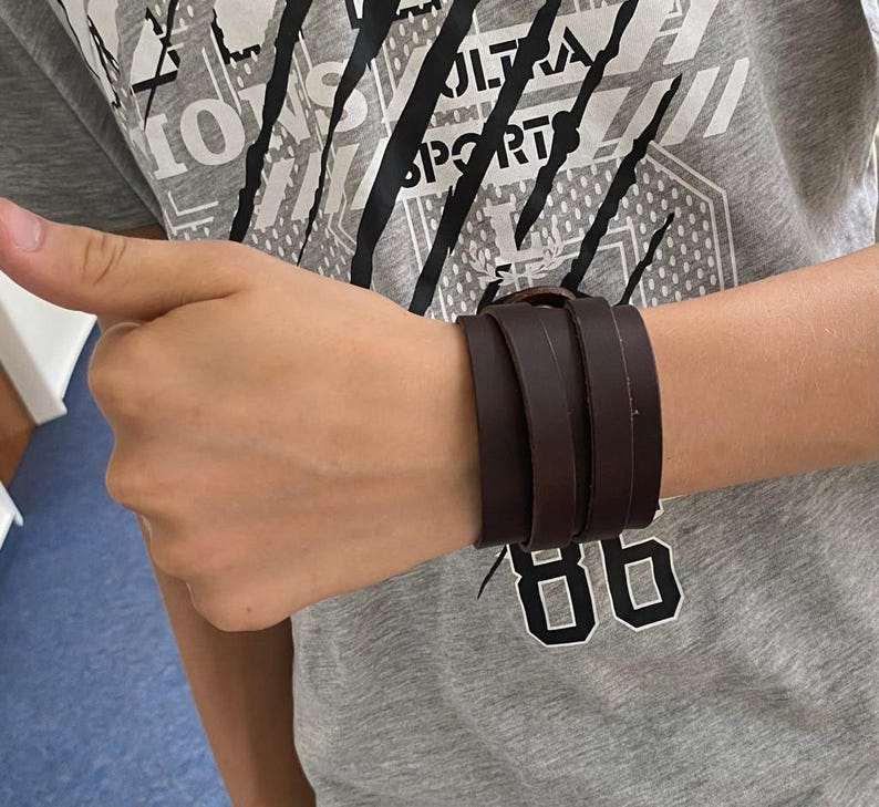 May include: A brown leather bracelet with three straps. The bracelet is worn on a person's wrist.