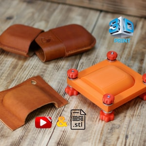 Könnte beinhalten: Ein offenes braunes Lederetui und ein passendes Lederstück werden auf einer Holzoberfläche gezeigt. Ein orangefarbenes quadratisches Gerät mit roten Knöpfen und dem Text "3D PRINT" sind ebenfalls sichtbar. Das Bild enthält auch ein Videosymbol und ein STL-Dateisymbol.