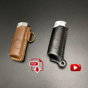 Könnte beinhalten: Zwei Leder-Feuerzeug-Etuis, eines braun und eines schwarz, jeweils mit einem genähten Design und einer Hunde-Silhouette. Das braune Etui hat einen goldenen Ring. Beide Etuis tragen den Aufdruck "HAND MADE".