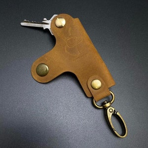 May include: A brown leather key holder with a brass snap closure and a brass key ring. The key holder has the word "HANDMADE" embossed on it.