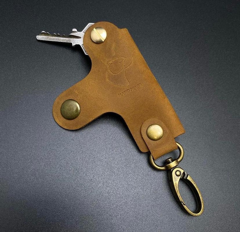 May include: A brown leather key holder with a brass snap closure and a brass key ring. The key holder has the word "HANDMADE" embossed on it.