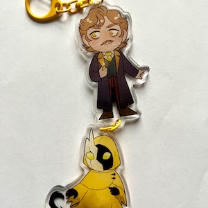 May include: A yellow and black character keychain with a yellow chain. The character is wearing a yellow hooded cloak and has a black and white face. The keychain also features a smaller character with brown hair, a mustache, and a purple coat.