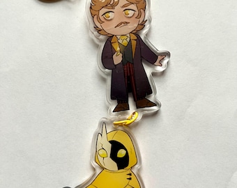 May include: A yellow and black character keychain with a yellow chain. The character is wearing a yellow hooded cloak and has a black and white face. The keychain also features a smaller character with brown hair, a mustache, and a purple coat.
