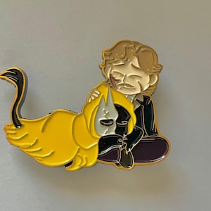 Puede incluir: Un pin de esmalte dorado con un personaje de dibujos animados con cabello castaño y un gato blanco y negro. El personaje lleva una chaqueta negra y pantalones morados. El gato lleva un disfraz amarillo y negro.